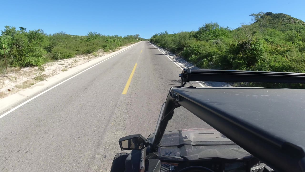Open road with yellow center line, vehicle hood, and low green vegetation.