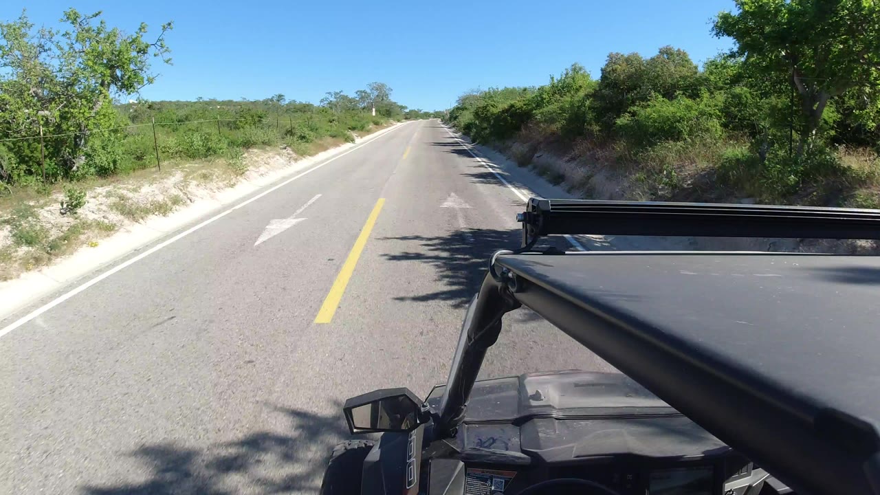 Vehicle-mounted view down a paved road in Los Cabos, Mexico, with green brush on both sides, a yellow center line, blue sky, and no visible people.