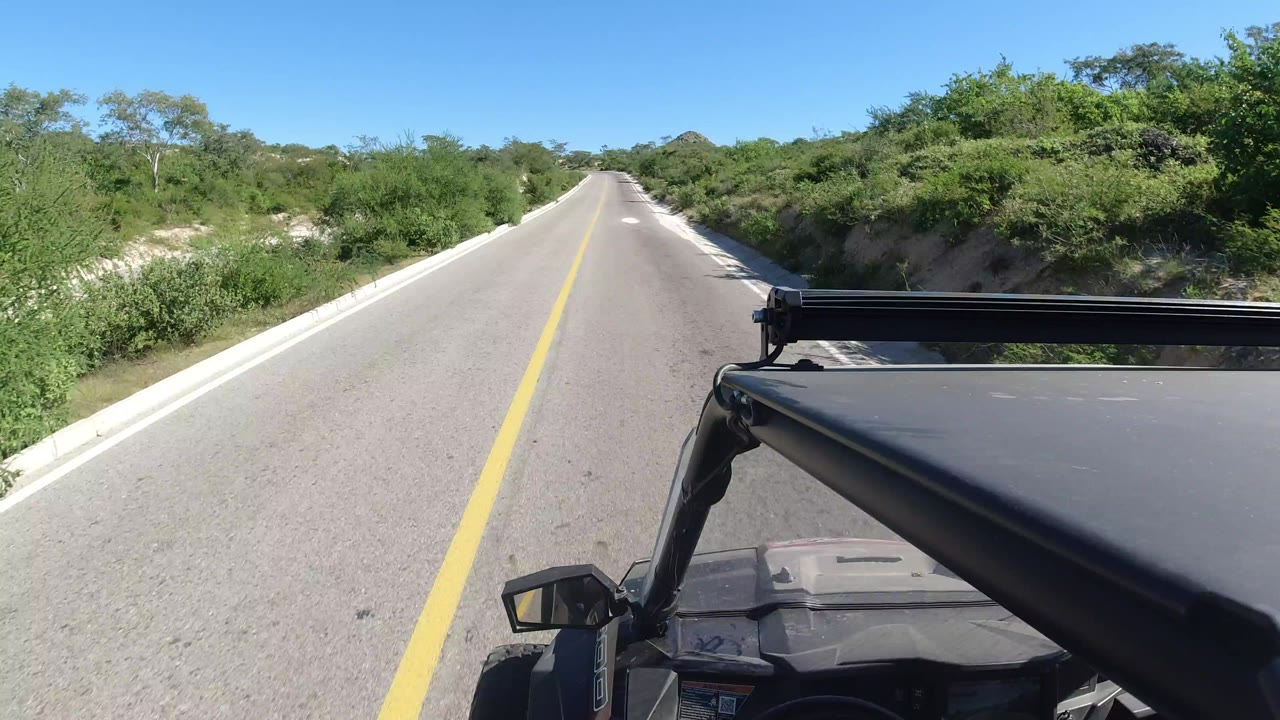 Forward vehicle view on a straight road in Los Cabos, Mexico, with a yellow center line, low green roadside vegetation, visible hood and roof rack, and no visible people.