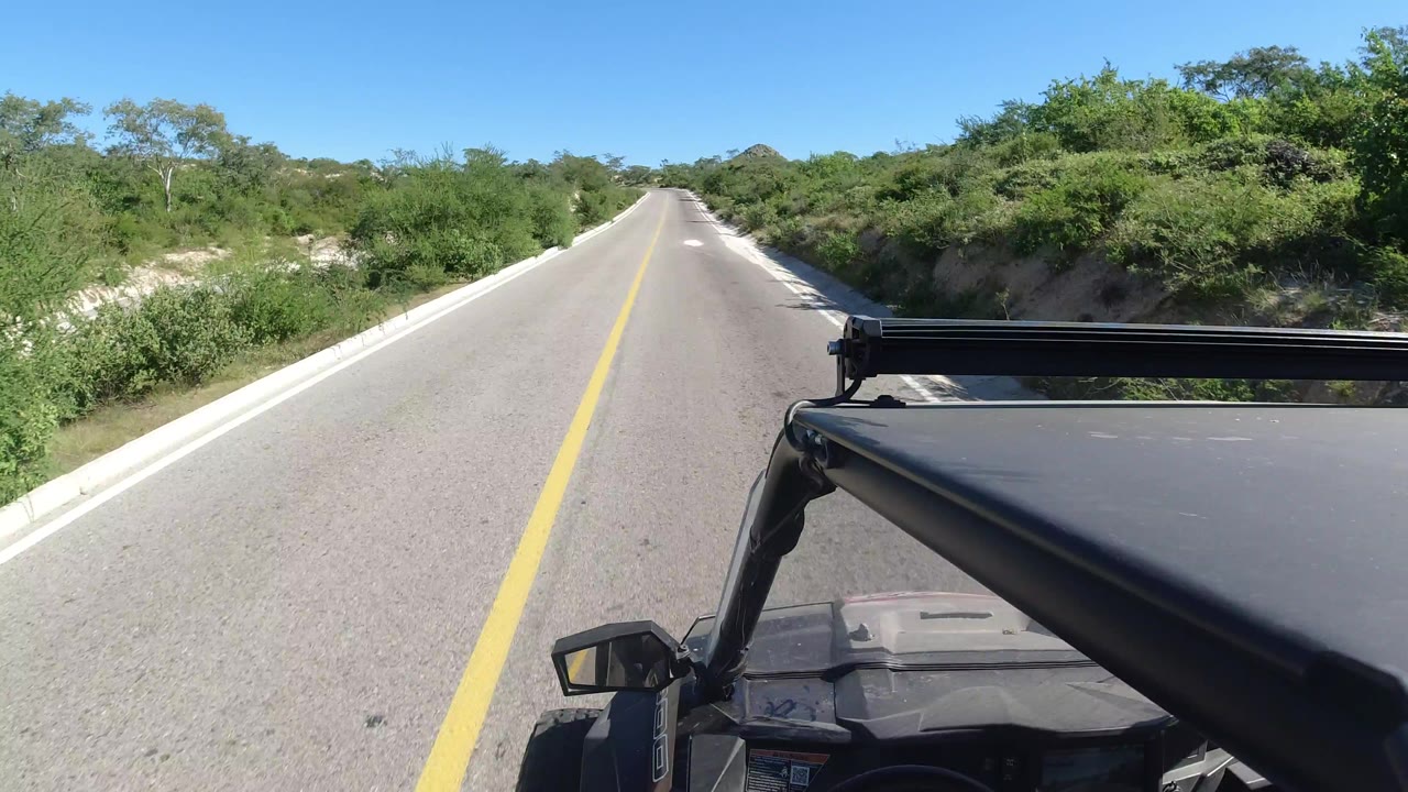 Vehicle POV along the same straight Los Cabos road, with the hood and black roof rack in the lower right, pale road shoulders, dense brush, and no visible people.