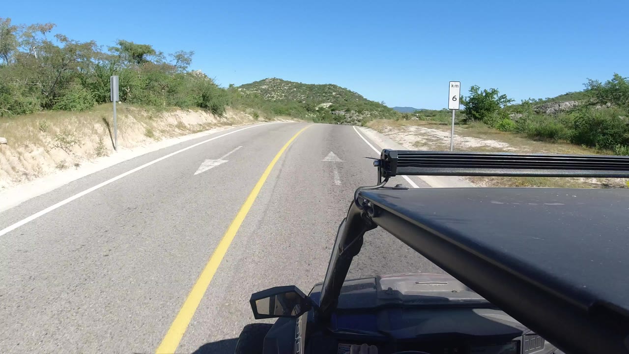 Jeep hood and light bar on a desert road with low hills ahead.