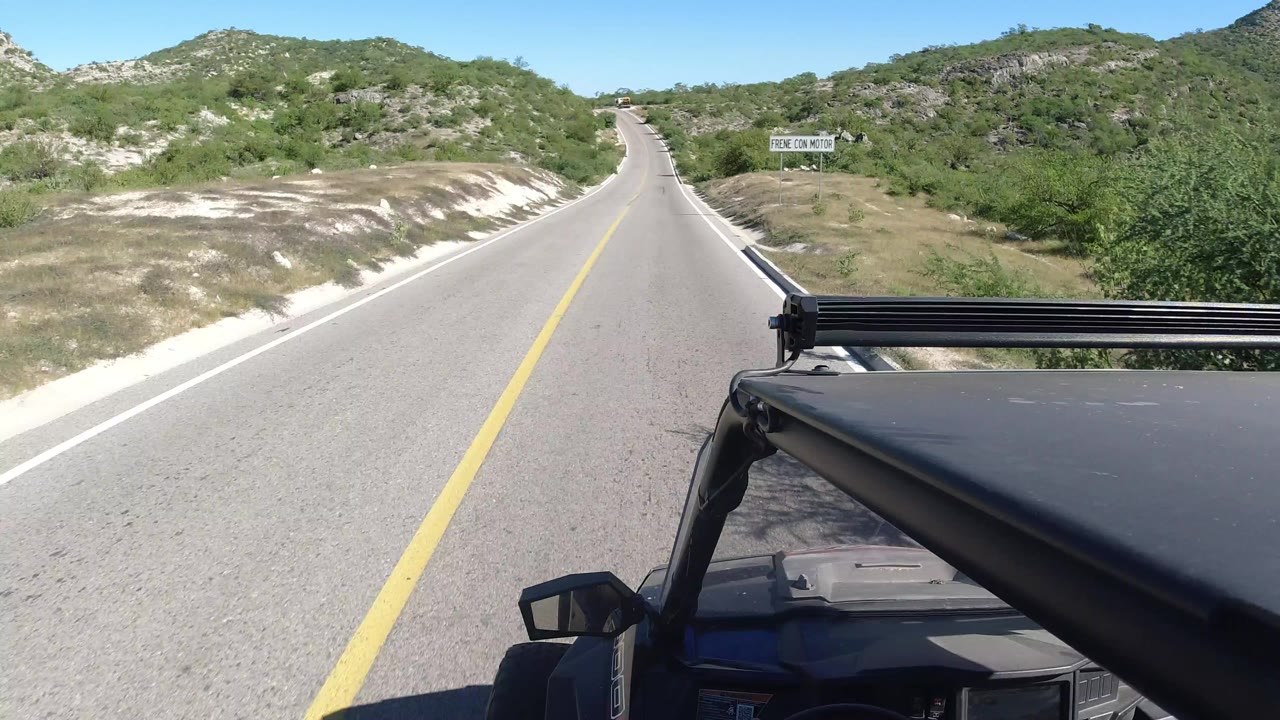 Opening frame of a forward road view in Los Cabos, Mexico, with a yellow center line, green hills, a roadside warning sign, the black vehicle roof in the foreground, and no visible people.