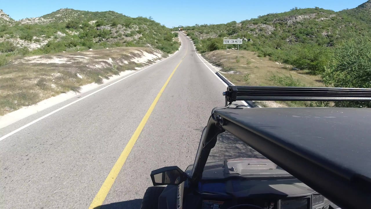 A vehicle-mounted view in Los Cabos, Mexico, looks up an empty two-lane highway bordered by pale shoulders, green brush, rocky slopes, and no visible people.