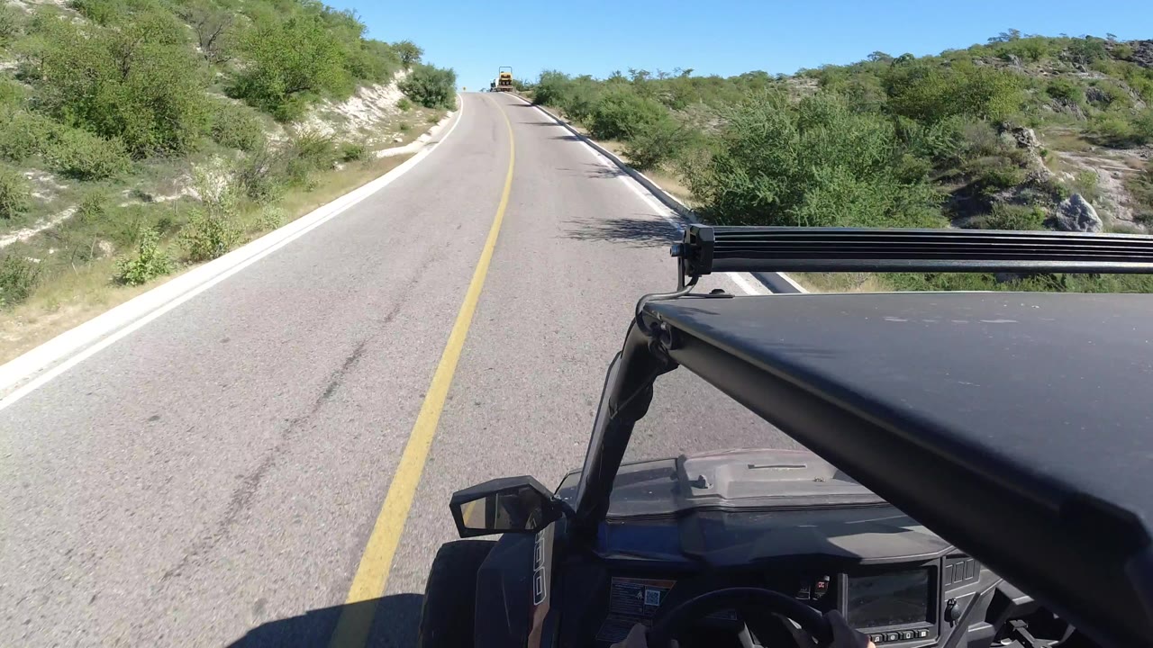 Vehicle-mounted view in Los Cabos looking down a straight paved road, with scrubby roadside vegetation, pale sand banks, and no visible people.
