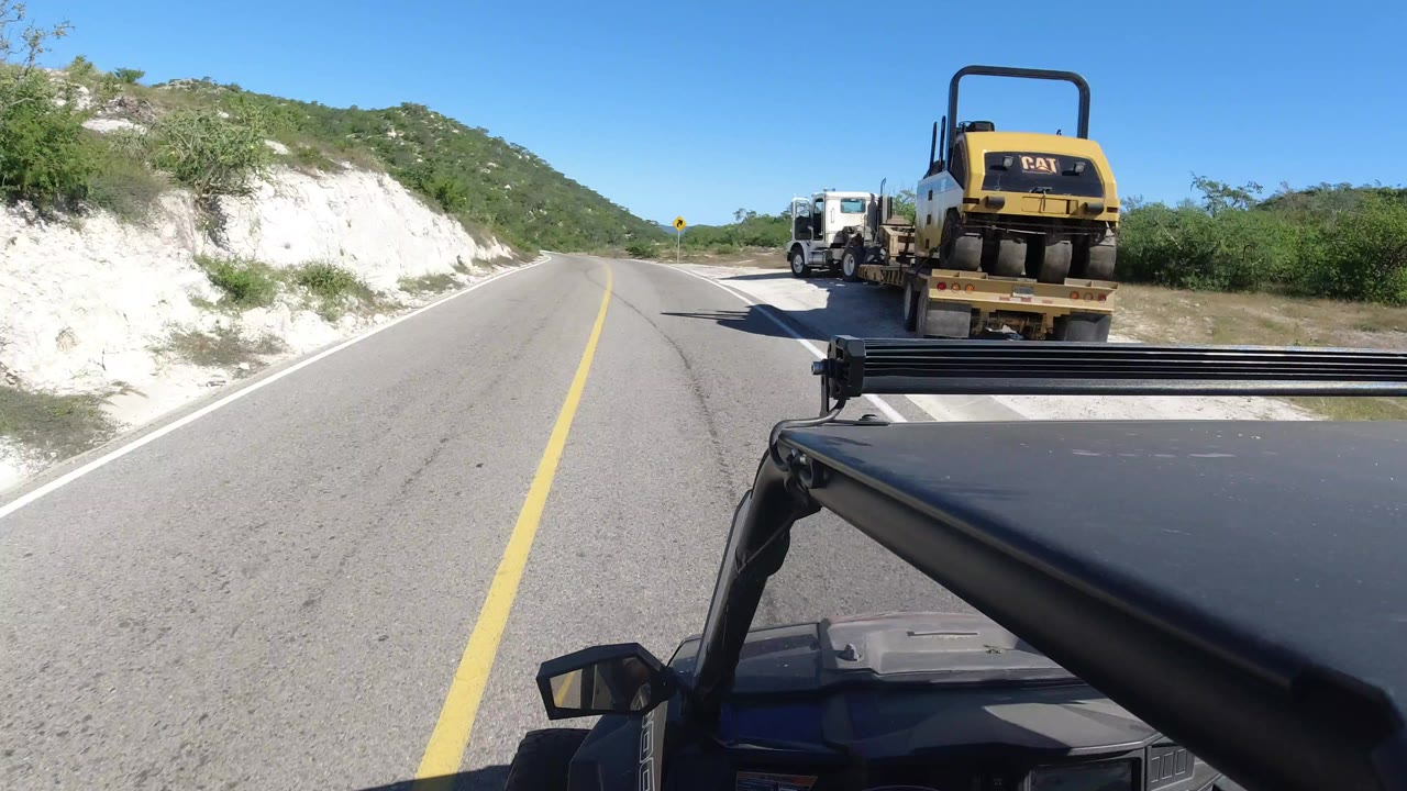 Forward vehicle view on a Los Cabos road with a flatbed truck and yellow road equipment ahead, scrub hills to the sides, bright blue sky, and no visible people.
