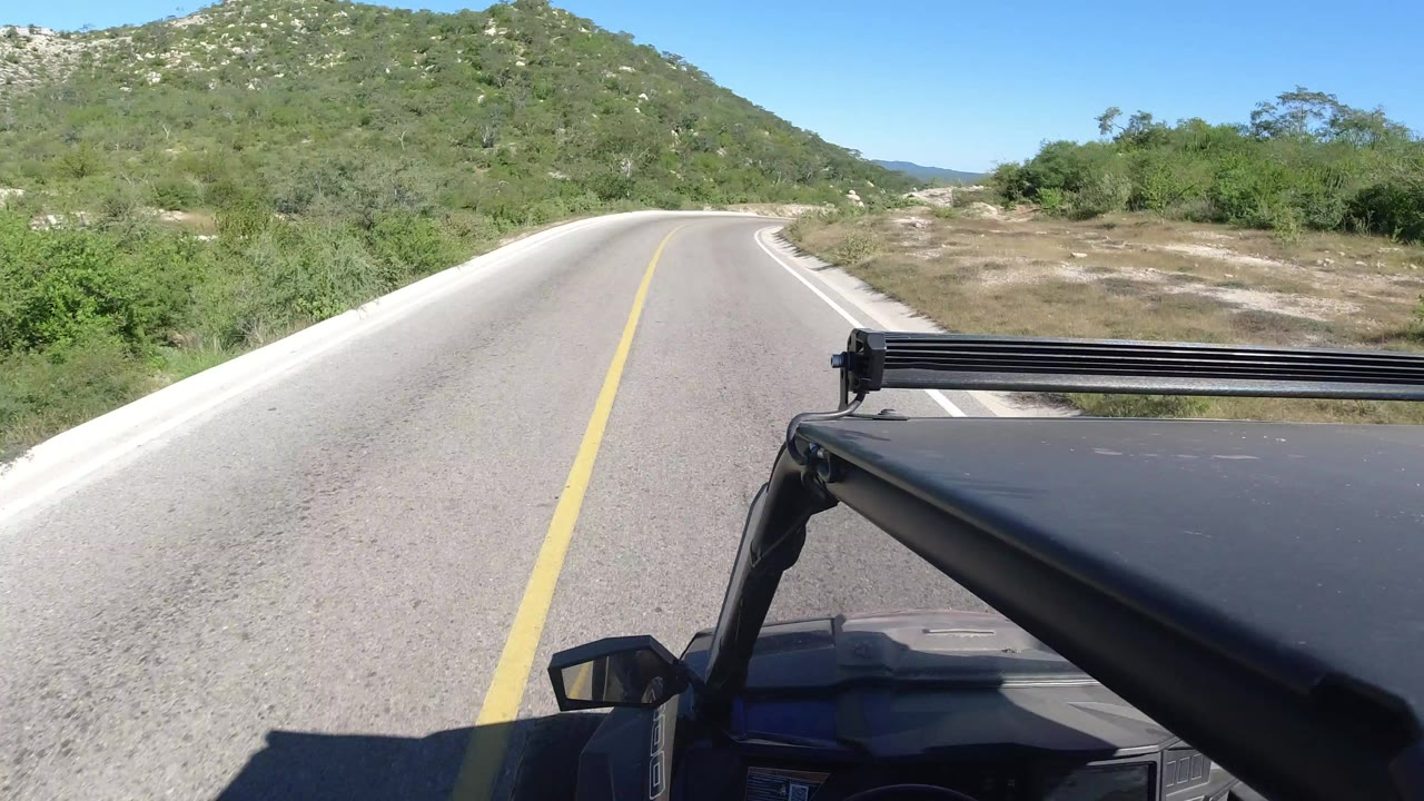 Forward vehicle view on a paved road curving through rocky green hills in Los Cabos, Mexico, with the hood and roof rack visible and no visible people.
