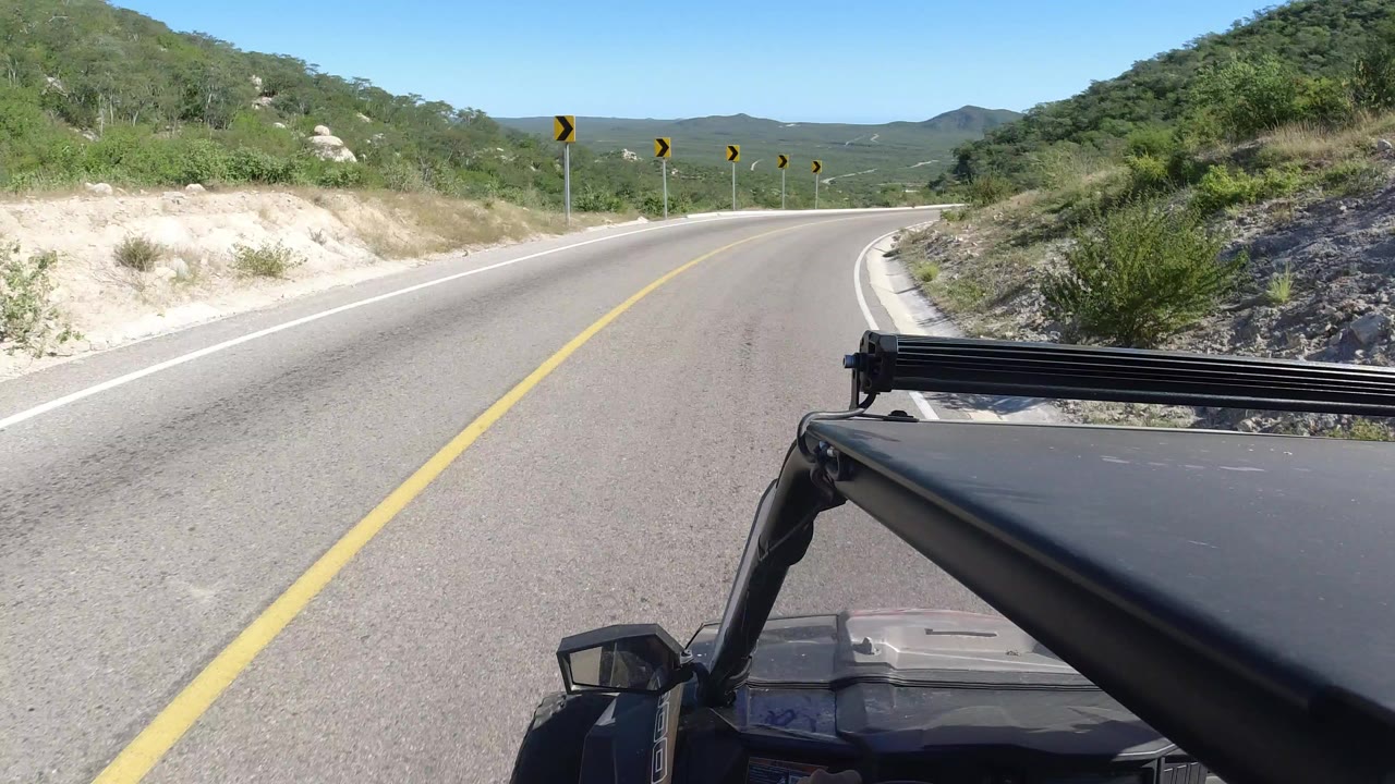 Opening frame of a Los Cabos two-lane highway curving right through green desert hills, with a vehicle roof edge and support bar visible in the foreground and no visible people.