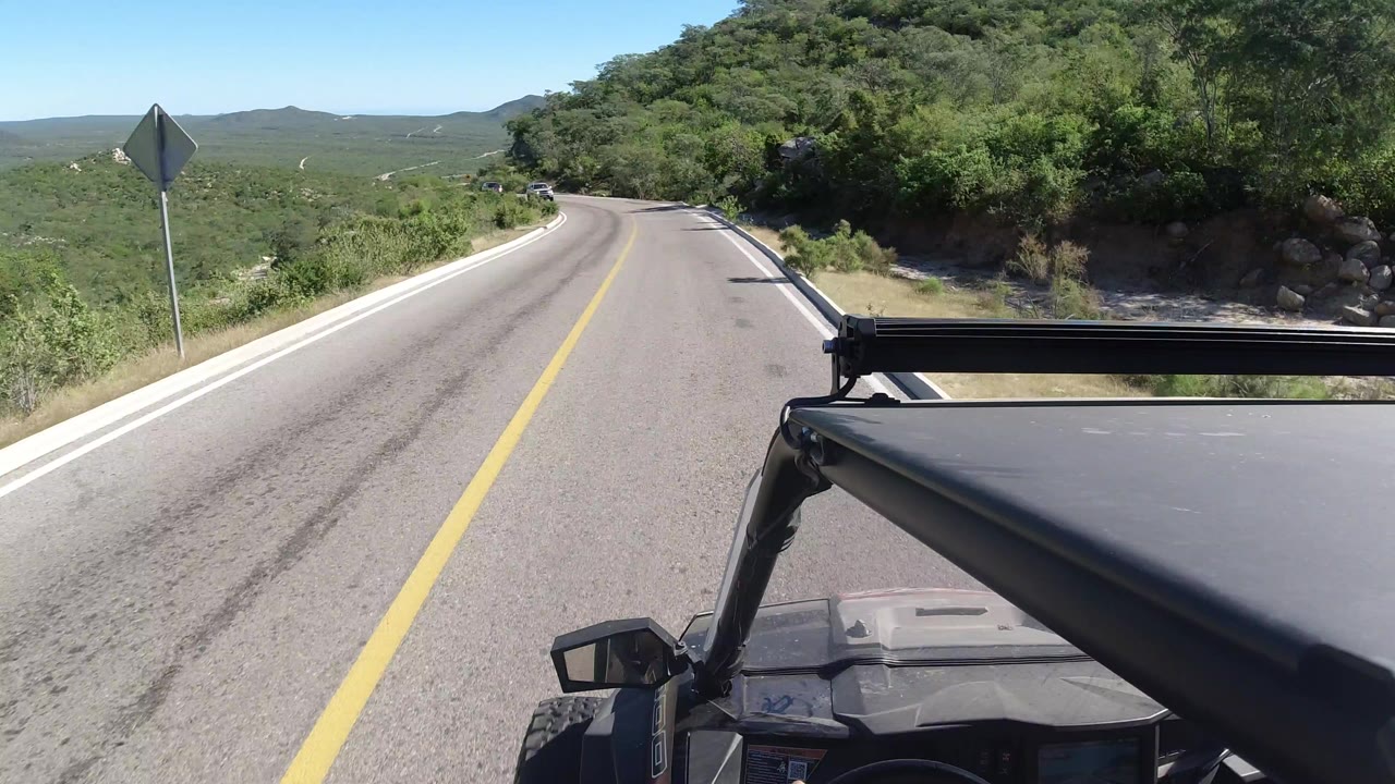 Two-lane road curving downhill with vehicle roof in foreground and green hills ahead.