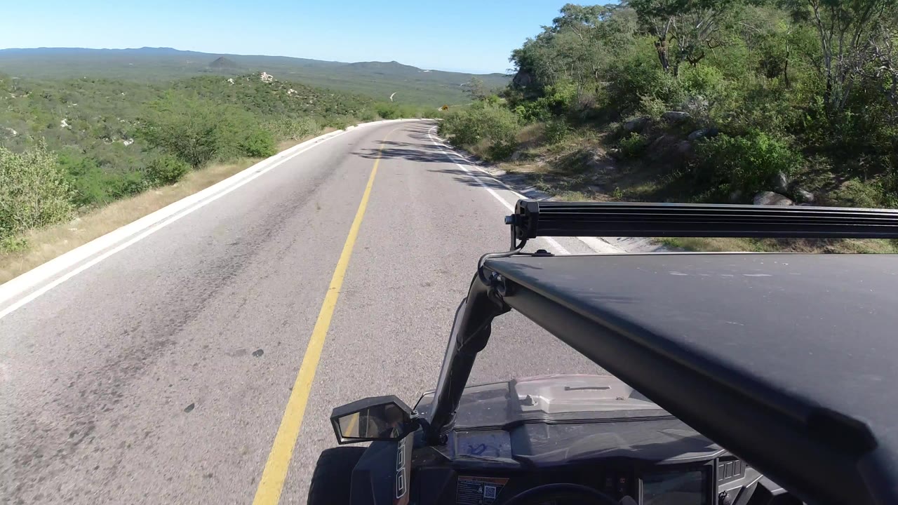 Forward view from an ATV on a rural road in Los Cabos, Mexico, with a yellow center line, green scrub hills, bright sky, and no visible people.