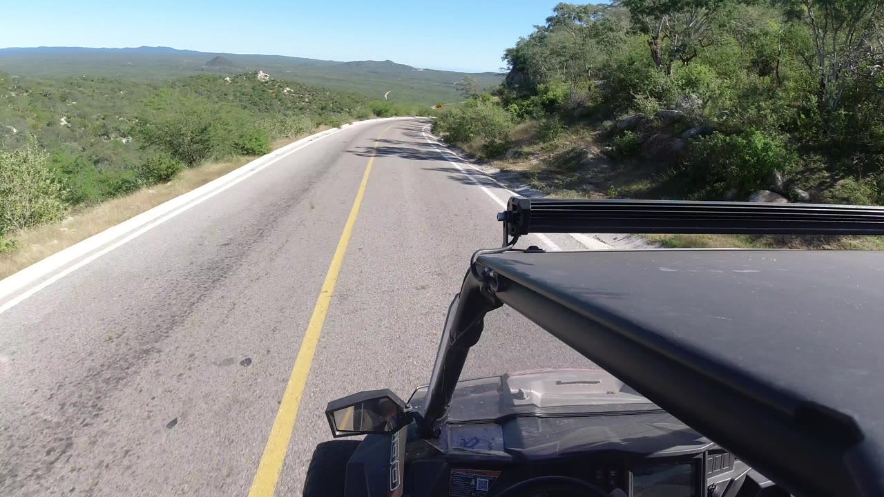 Vehicle-mounted view on a sunlit rural road in Los Cabos, Mexico, with the ATV roof at lower right, roadside brush, distant hills, and no visible people.