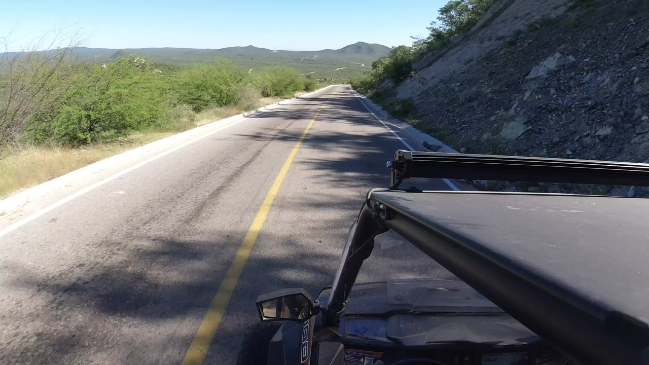 Opening view from a vehicle on a narrow paved road in Los Cabos, Mexico, with a yellow center line, roadside brush, a rocky slope at right, distant hills, and no visible people.