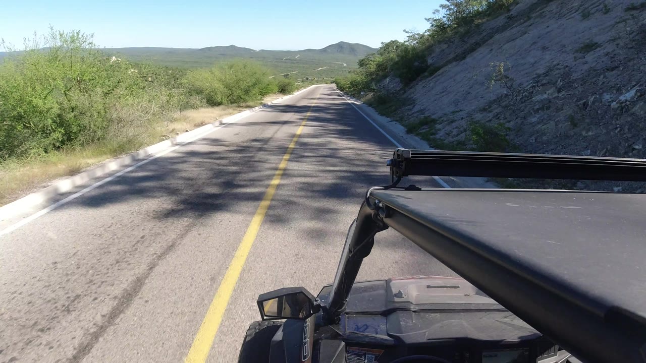 Vehicle POV passing a shaded section of narrow road near a rocky cut.