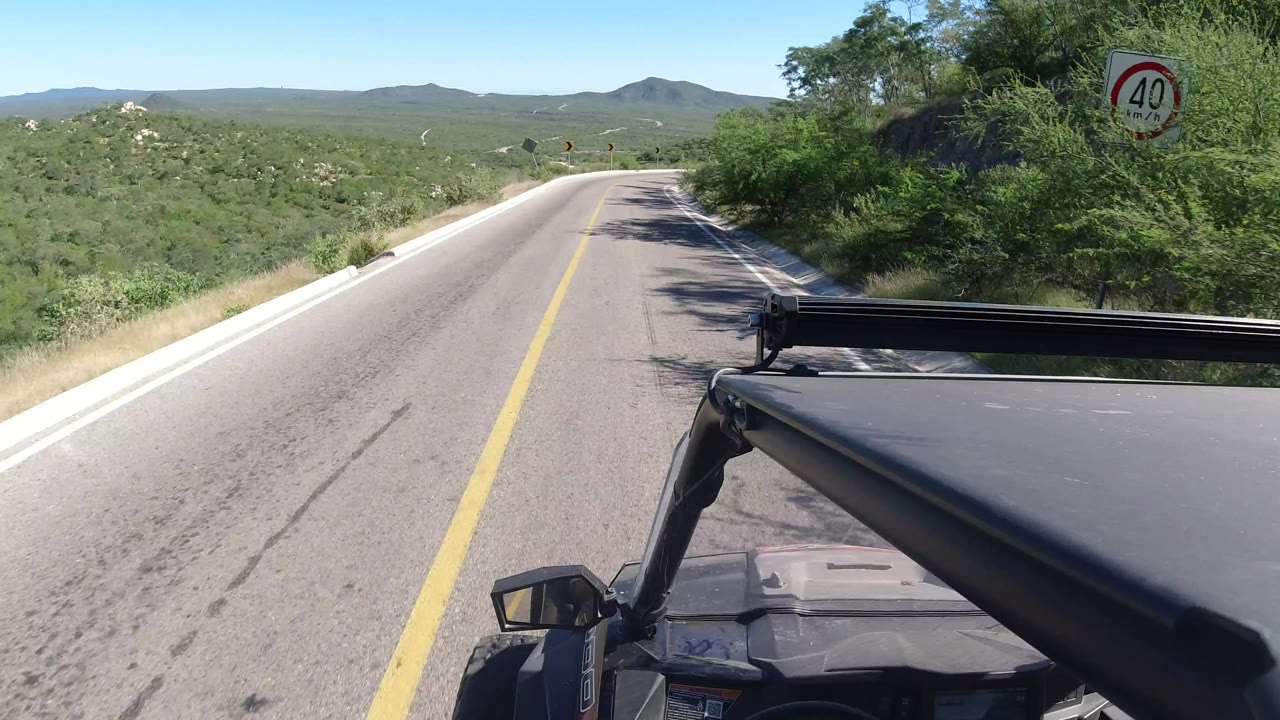 Forward view from a jeep on a sunny Los Cabos, Mexico road, with a 40 speed sign at right, green roadside brush, distant hills, and no visible people.