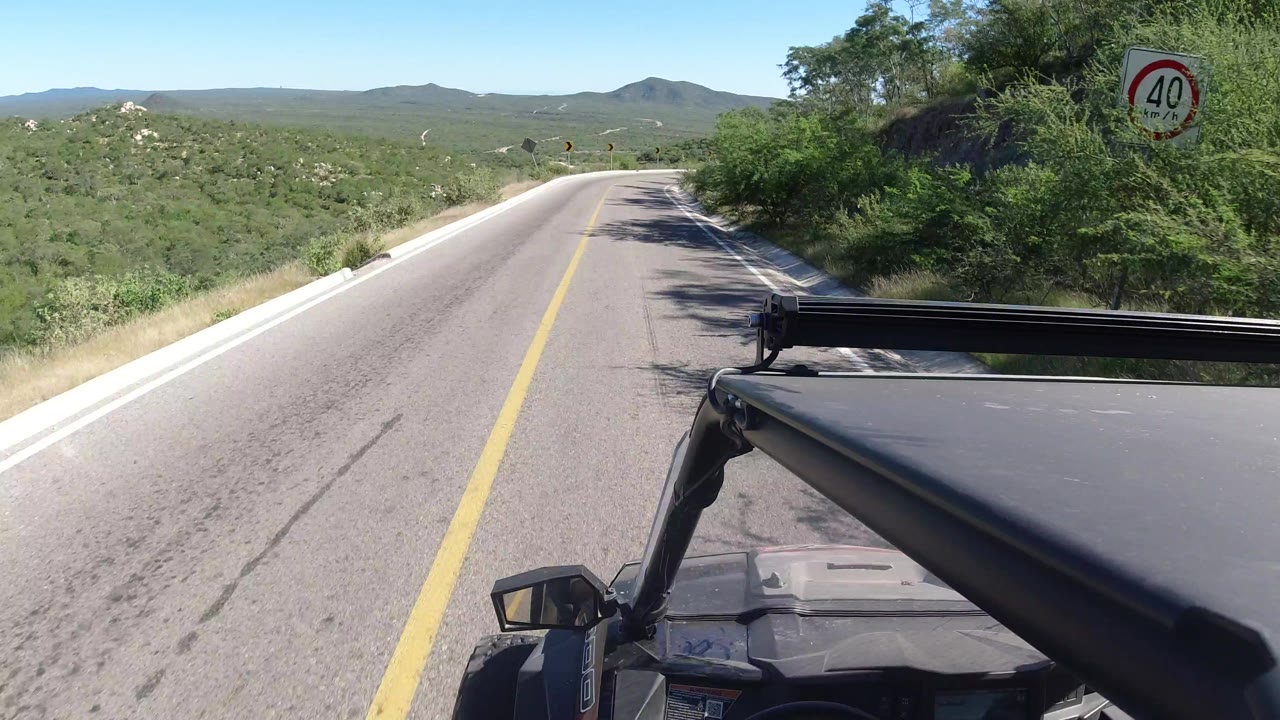 The jeep continues along an empty Los Cabos road with yellow center line, low mountains ahead, dense green brush at right, and no visible people.