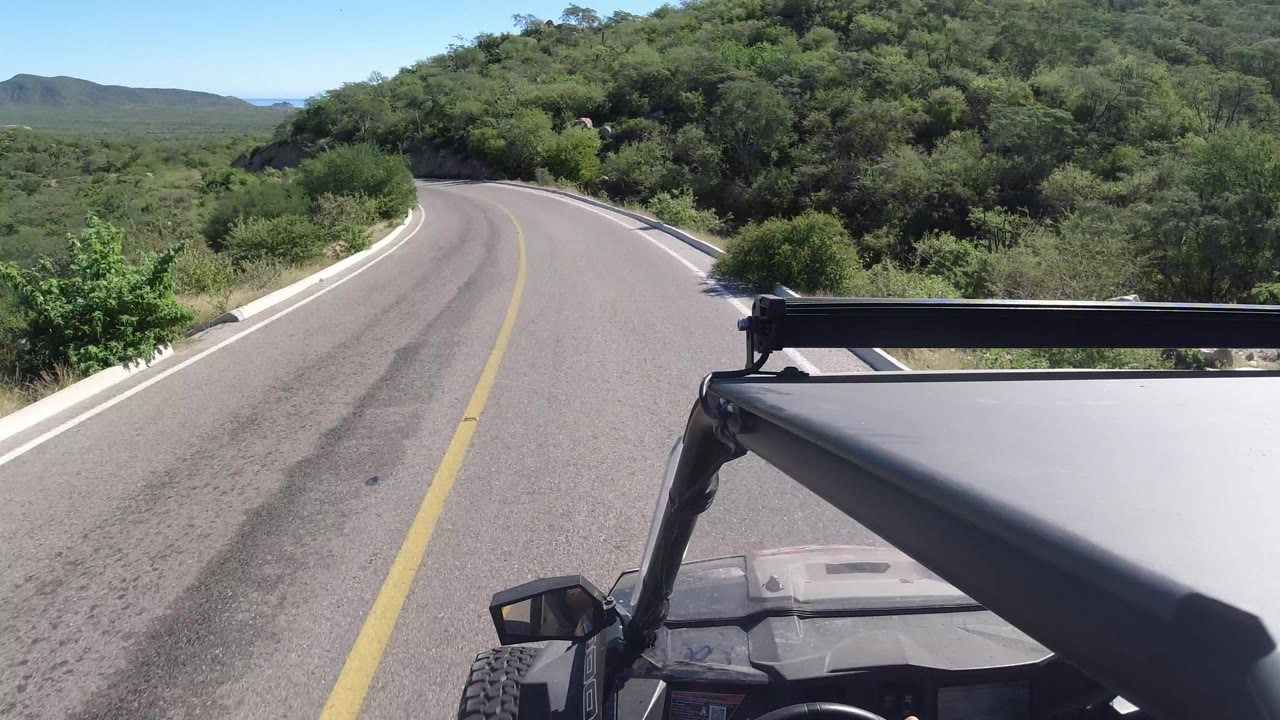 Los Cabos road view from an open vehicle as a two-lane paved road curves left through dense green vegetation under a clear blue sky, with no visible people.