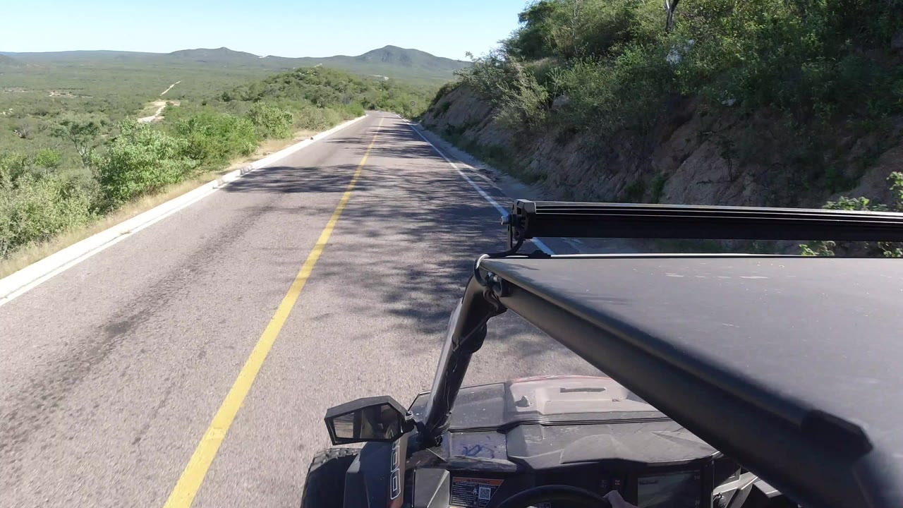 Forward-facing view from an open vehicle on a narrow road in Los Cabos, Mexico, with green roadside brush, distant hills, and no visible people.