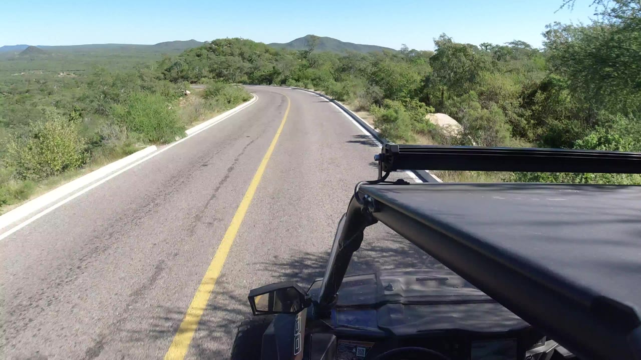 Curving paved road seen from a moving vehicle beside green scrubland.
