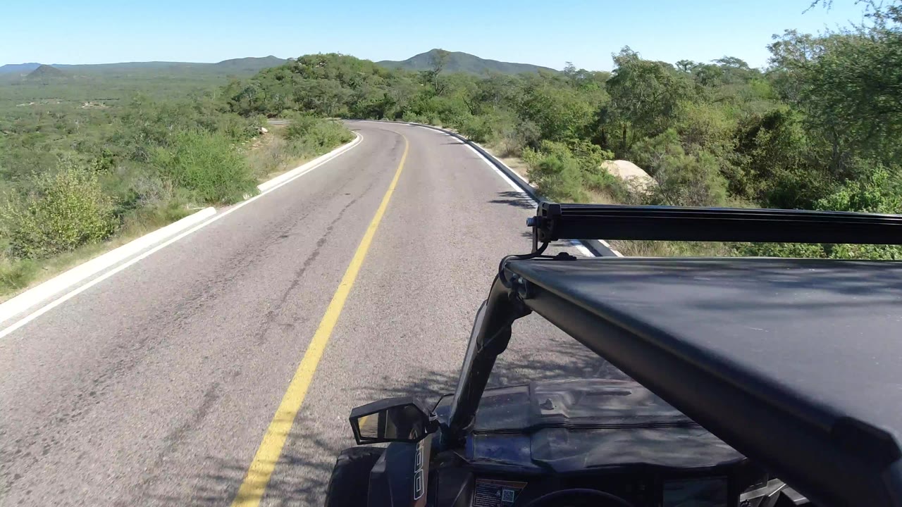 A Los Cabos roadway stretches ahead with a yellow center line, brush on both sides, distant hills, and the vehicle structure filling the lower right foreground.