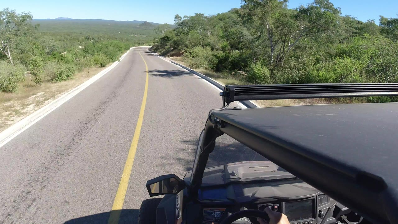 The Los Cabos road continues straight downhill, framed by dense green roadside brush, a black off-road vehicle roof at lower right, distant low mountains, and no visible people.