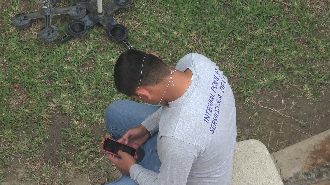 At 0:00 in Los Cabos, Mexico, a man in a gray shirt sits near grass and a concrete edge while holding a phone with a red case.