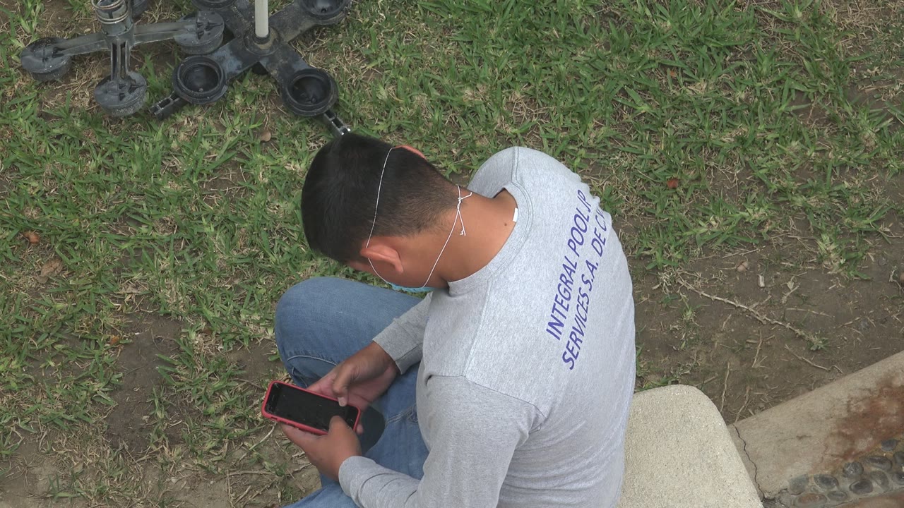 At 0:01 in Los Cabos, Mexico, the seated worker remains angled away from camera, looking down at the phone in his hands.