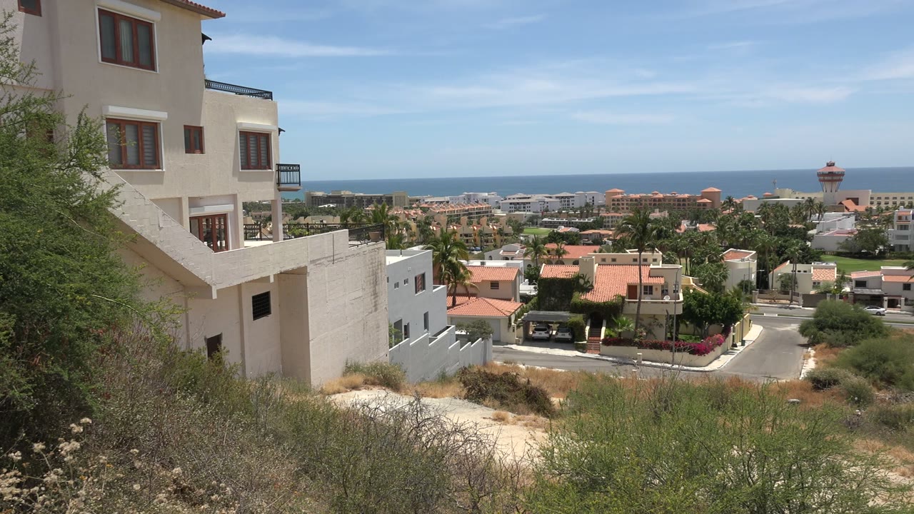 Opening frame of Los Cabos hillside homes with dry scrub in front, red tile roofs below, blue ocean on the horizon, and no visible people.