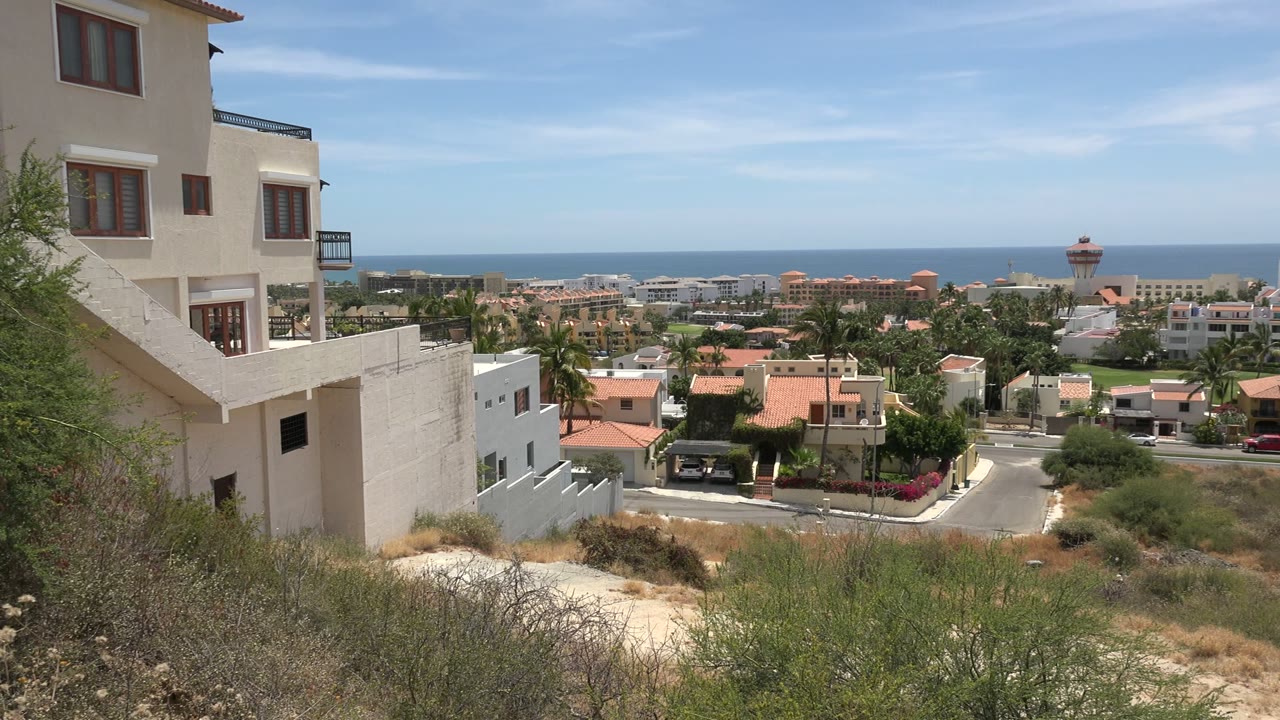 Los Cabos neighborhood view from a hillside position, with a cream building at left, palm trees and red roof homes below, ocean beyond, and no visible people.