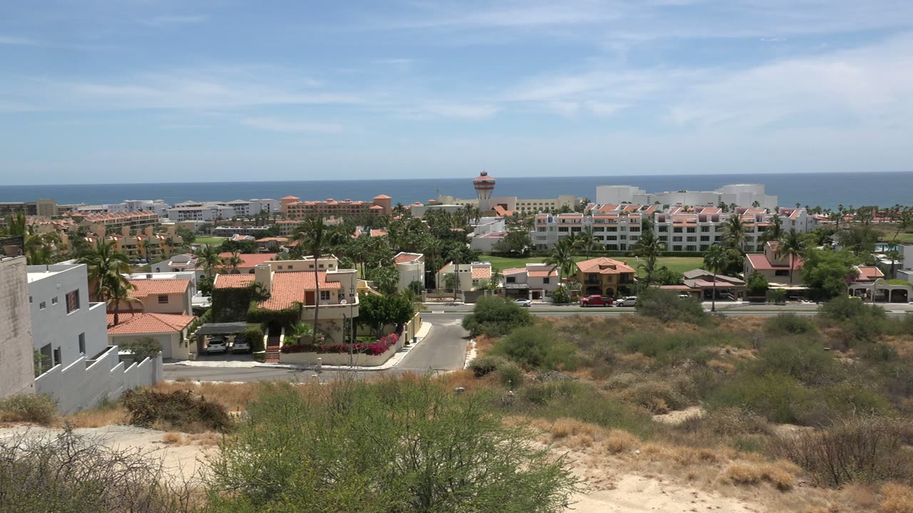 Early pan view across Los Cabos with sandy brush in front, a roadside neighborhood or resort area in the middle distance, ocean beyond, and no visible people.