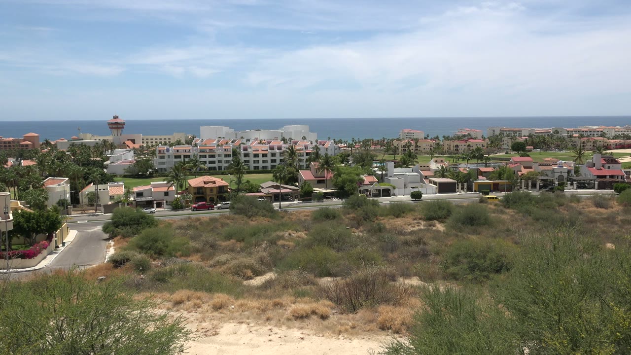 Los Cabos, Mexico frame with scrubby sand in front, a road and low coastal buildings in the middle distance, and a flat blue ocean horizon under thin clouds, with no visible people.