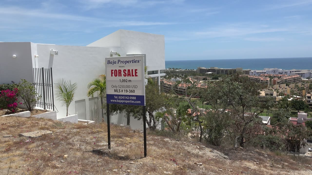 Opening frame in Los Cabos, Mexico, with a Baja Properties for sale sign near a white building, dry hillside ground, ocean horizon, and no visible people.