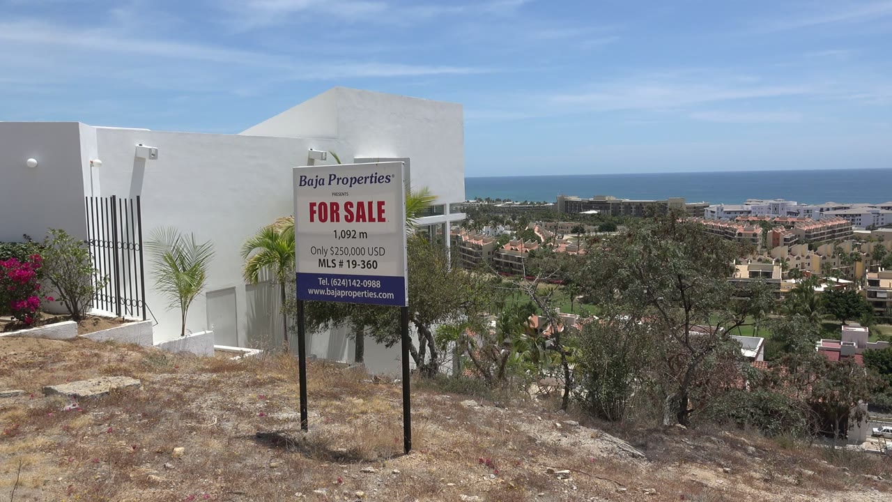 Second frame shows the sale board more centered against the white Los Cabos building, with hillside brush, distant rooftops, sea, and no visible people.