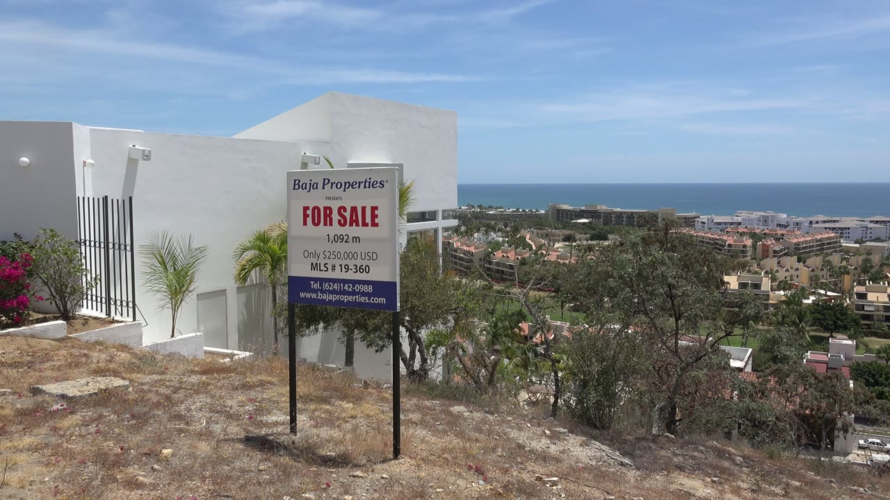 For-sale sign on a dry hillside with coastal buildings and ocean beyond.