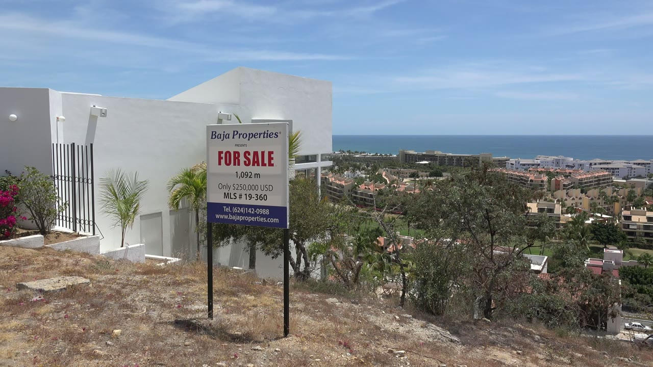 At 0:01, the Los Cabos frame keeps the for-sale sign near the white wall, with palms at left, hillside vegetation below, clustered buildings in the distance, ocean, and no visible people.