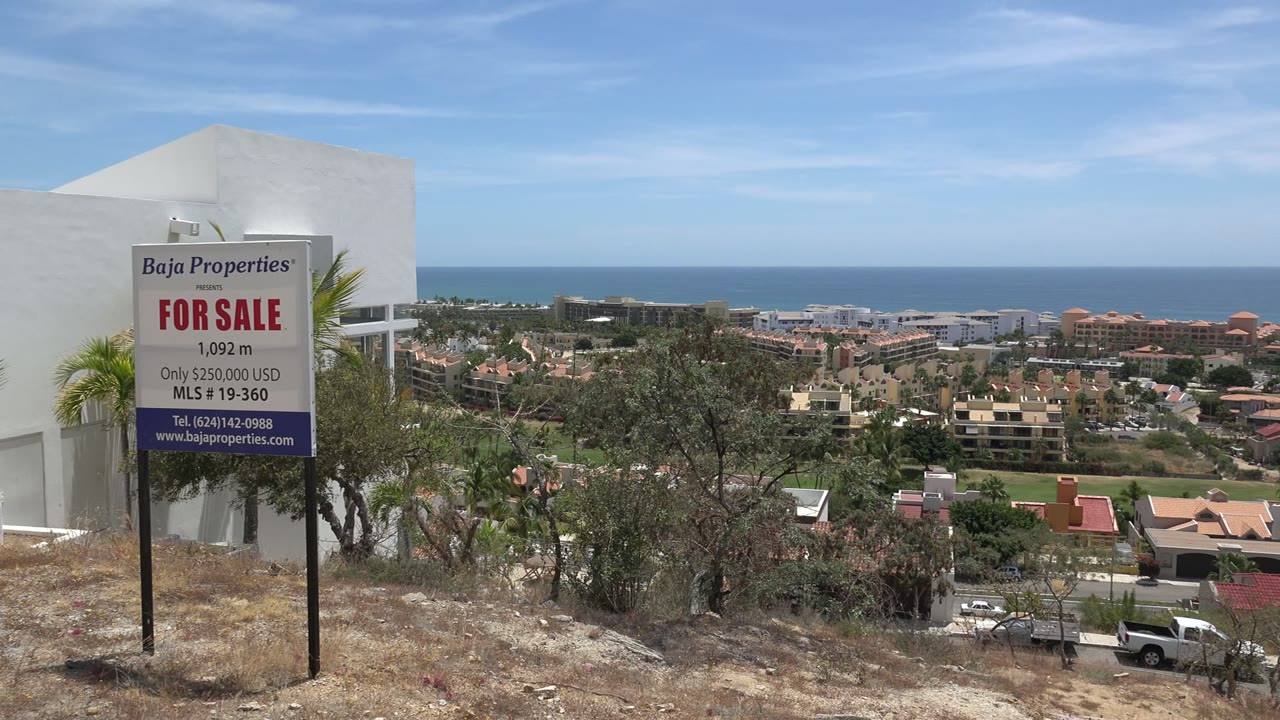 For-sale sign on a dry hillside above houses and the ocean
