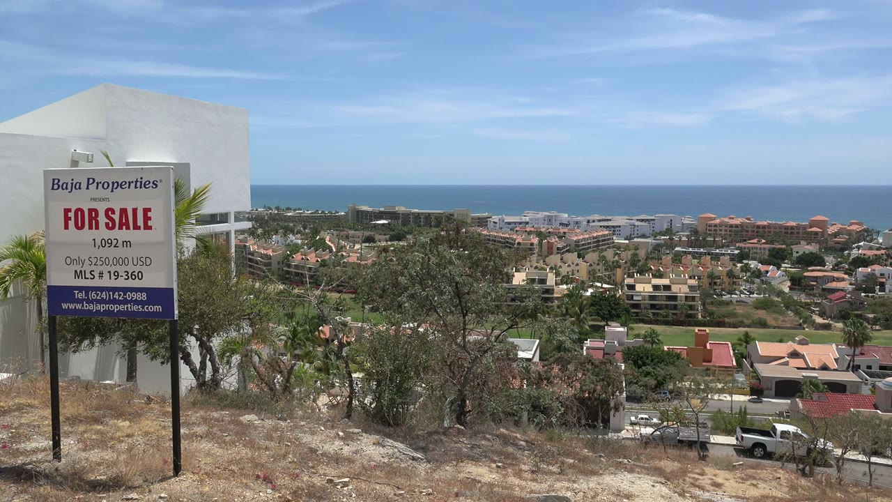 Los Cabos overlook with the for-sale sign still prominent, scrubby foreground, a white truck on the road below, layered houses, ocean, and no visible people.