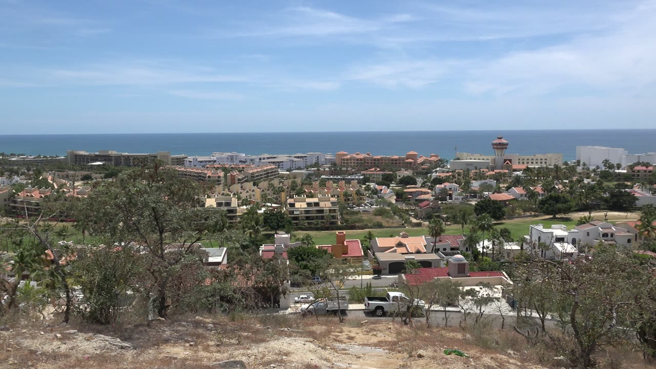 Wide hillside view over a Los Cabos residential area with dry brush in front, homes and trees below, ocean on the horizon, and no visible people.