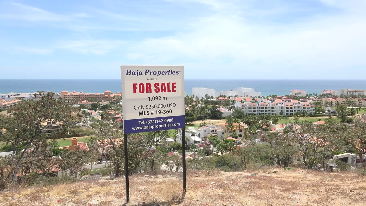 For sale sign on a hillside above coastal buildings and ocean.