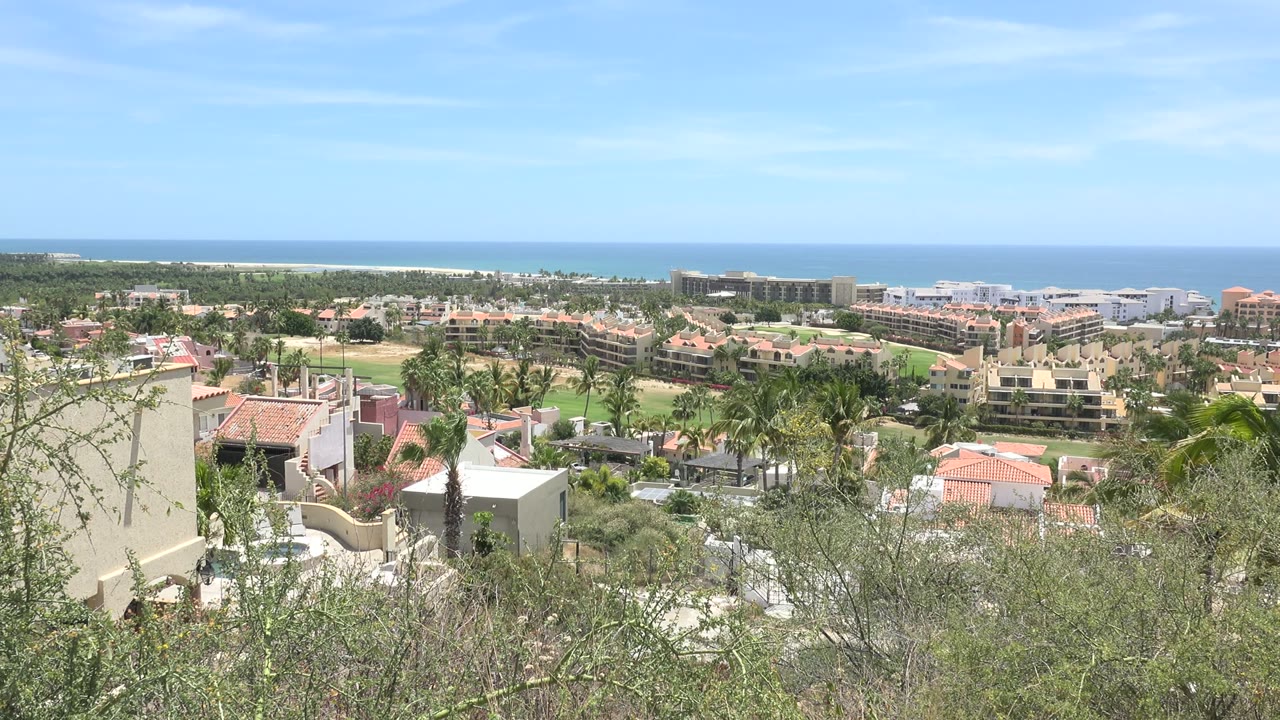 One second into the Los Cabos, Mexico clip, the wide frame holds on palms, green grounds, red roofs, pale buildings, blue ocean, and no visible people.