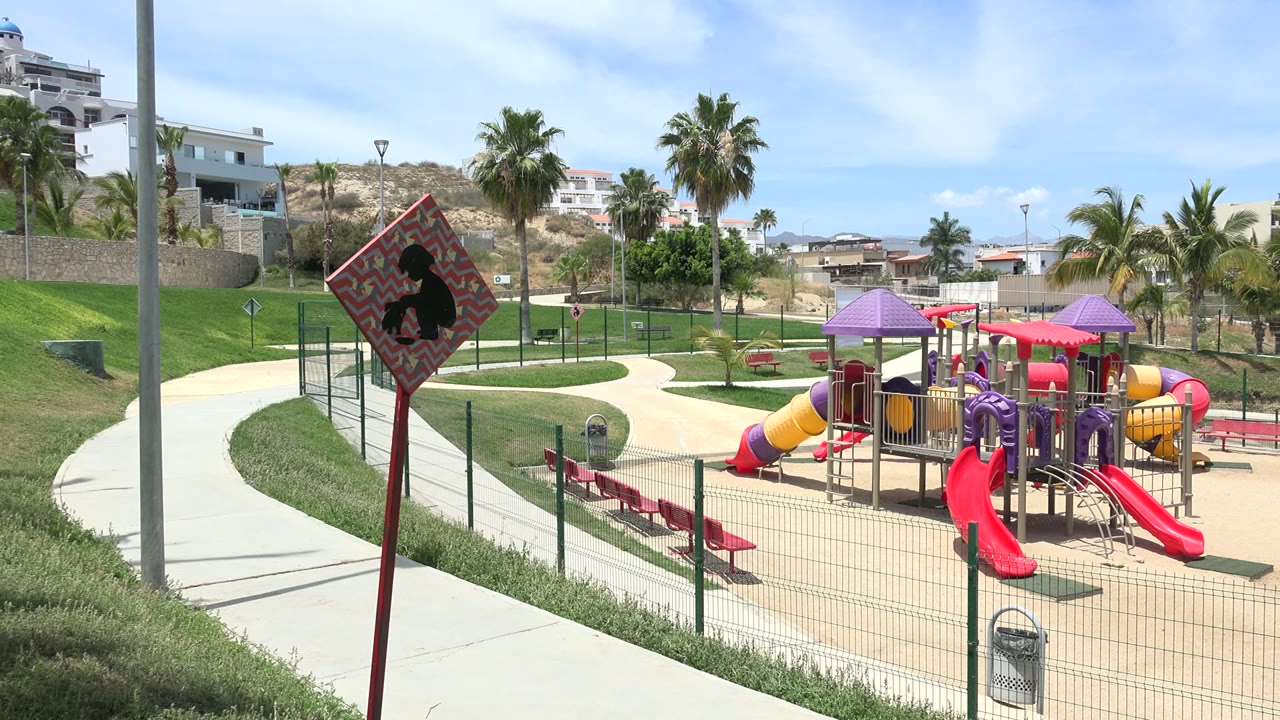 Opening frame of a Los Cabos playground with red slides, purple roof panels, a foreground sidewalk, palms, hillside buildings, and no visible people.