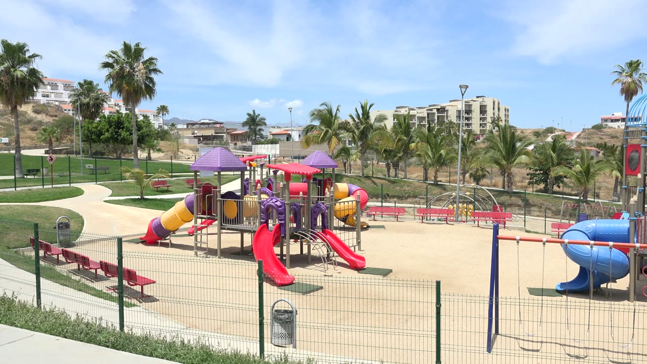 Wide view of a colorful fenced playground with palm trees and benches.