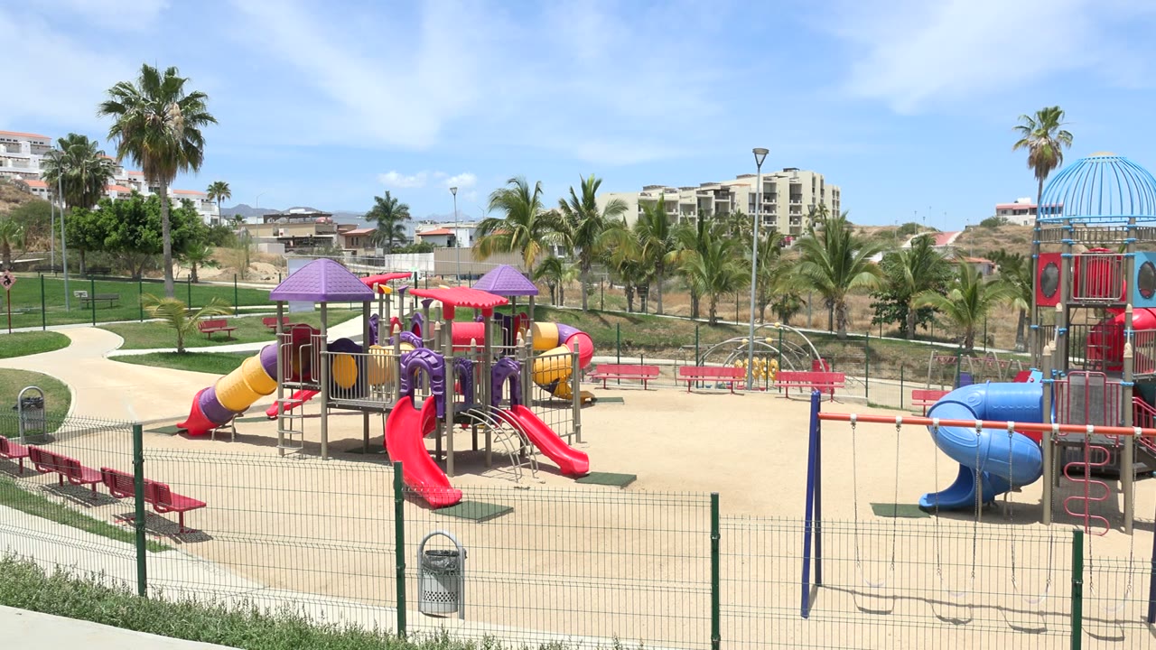 At 0:01, the frame centers the Los Cabos play area with twin red slides, a swing set in front, pink benches to the left, palms behind, and no visible people.