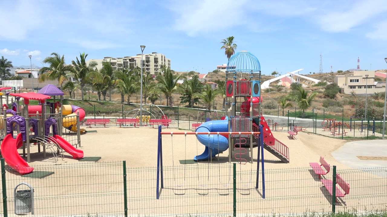 Wide sunny playground with slides, swings, benches, palms, and a fence.