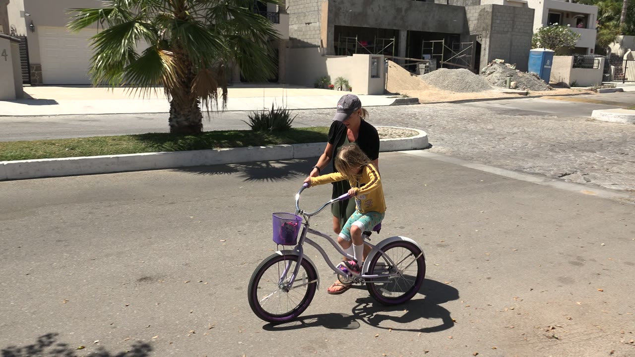 In Los Cabos, a woman in dark clothing walks beside a child in a yellow top riding a purple bicycle across a sunlit empty street.