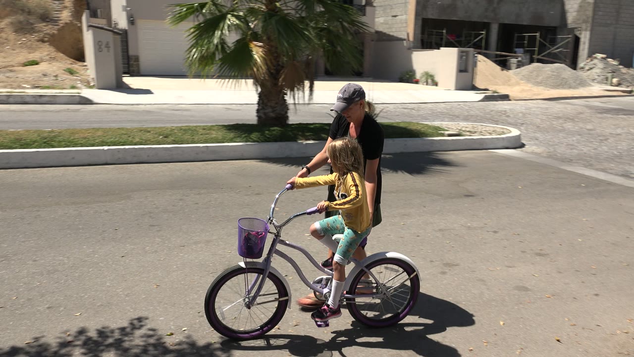 The child pedals the purple bicycle farther along the Los Cabos street while the woman stays close at her side under strong daylight.