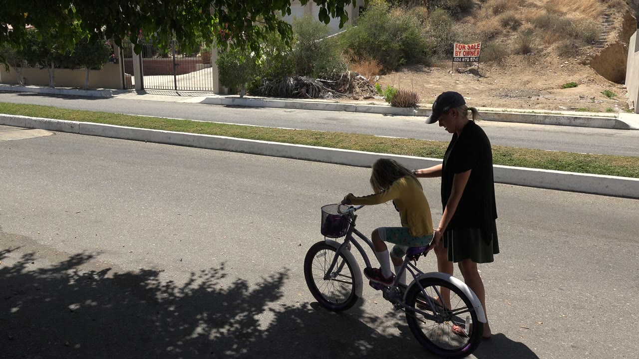 Adult woman steadies a child on a bicycle in a shaded street.