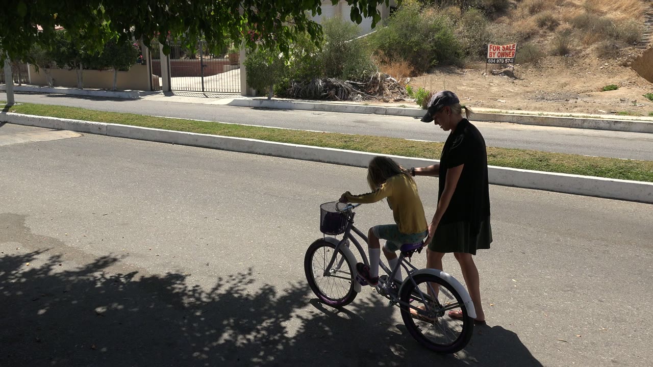 In Los Cabos, Mexico, the child moves slightly forward on the bicycle as the adult woman walks behind, with a curb, grass strip, gate, and dry hillside across the road.