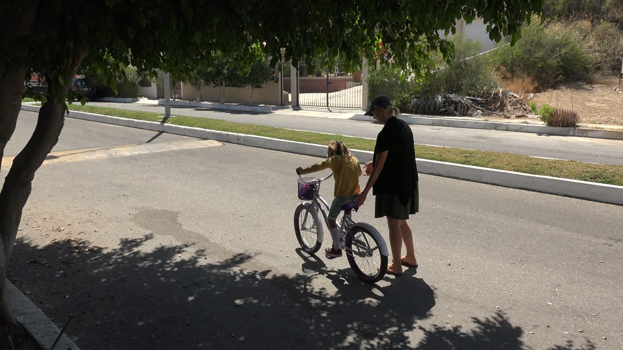 In Los Cabos, Mexico, a woman stands beside a young girl on a small bicycle under tree shade at the start of the clip.