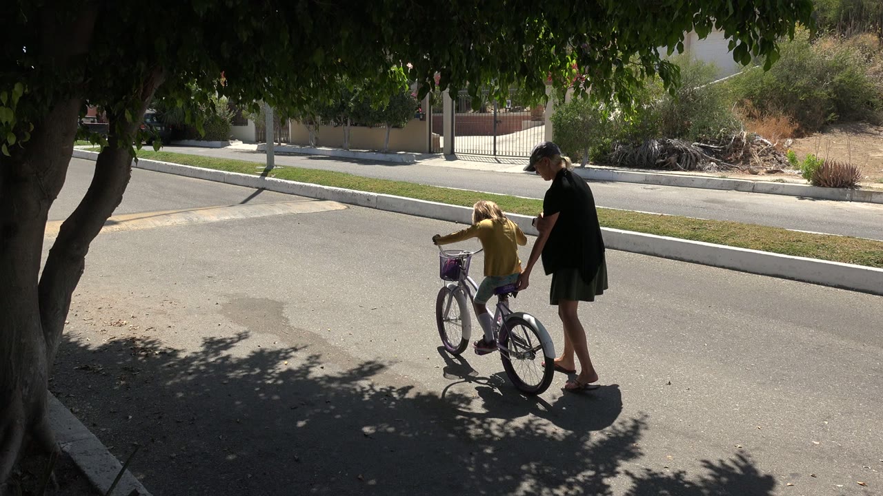 In Los Cabos, Mexico, the girl holds the handlebars while the woman stays close beside the bicycle on the sunlit road.