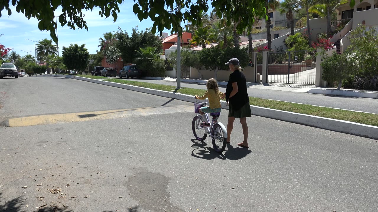 Girl on a bicycle beside an adult woman on a sunny residential street.