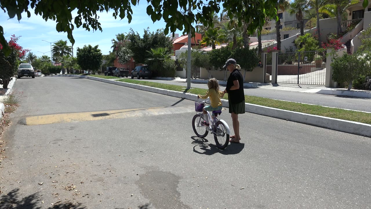 The girl remains on the bicycle as the adult woman stays close beside her on the sunlit Los Cabos residential road, with shade from leaves across the top of frame.